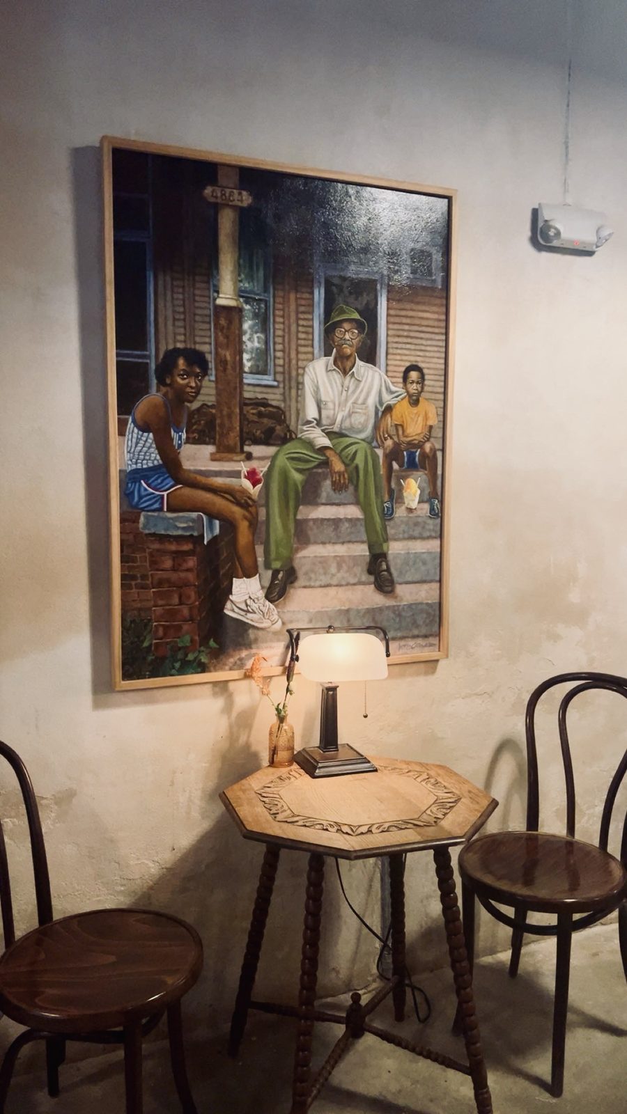 A cozy café corner with a carved wooden table, a lamp and a large painting of a family on porch steps